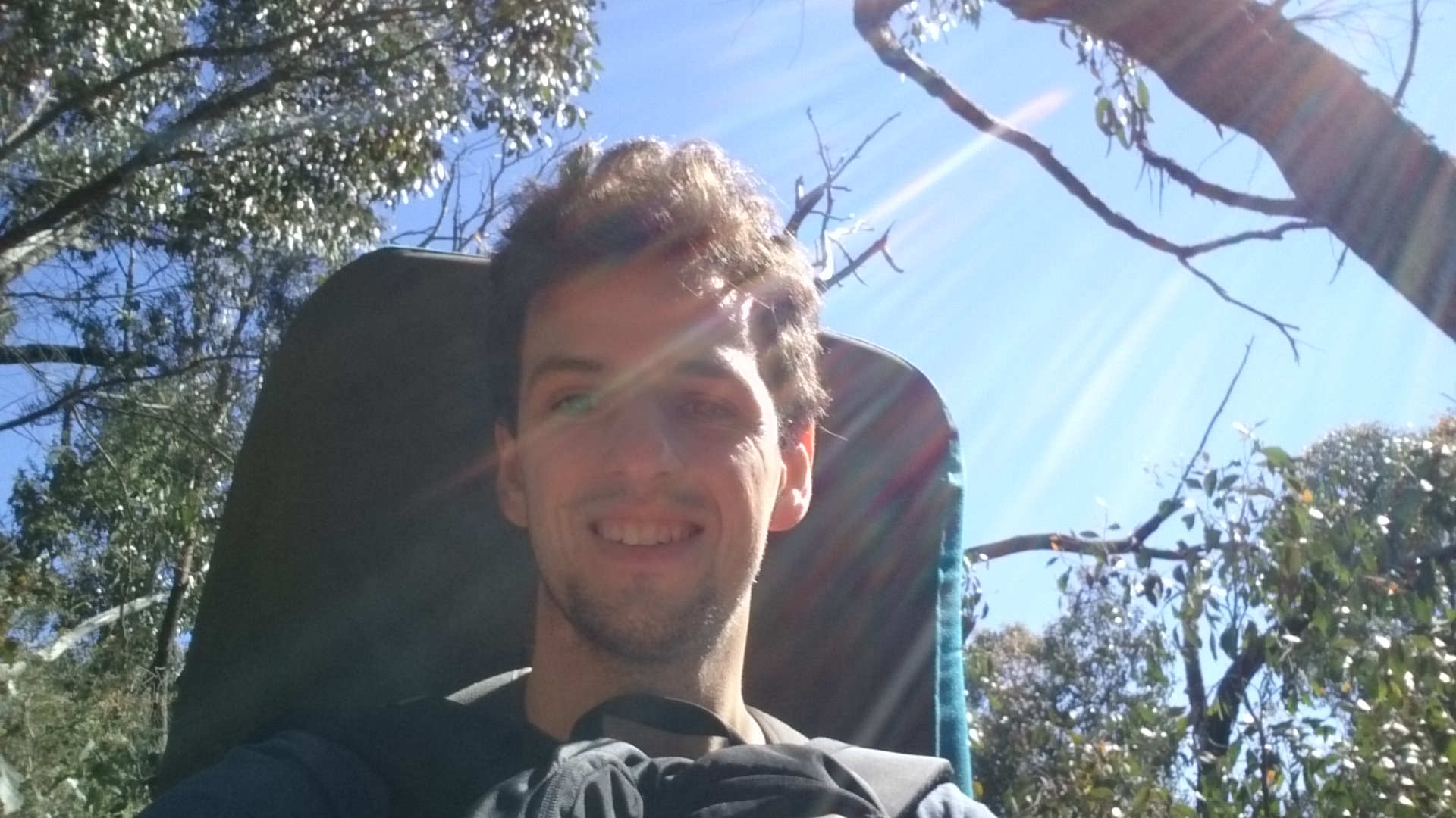 Oliver Robson looking at the camera while wearing a bouldering mat on his back, a backpack on his front, walking through a sparse Australian forest while the sun shine down on the camera